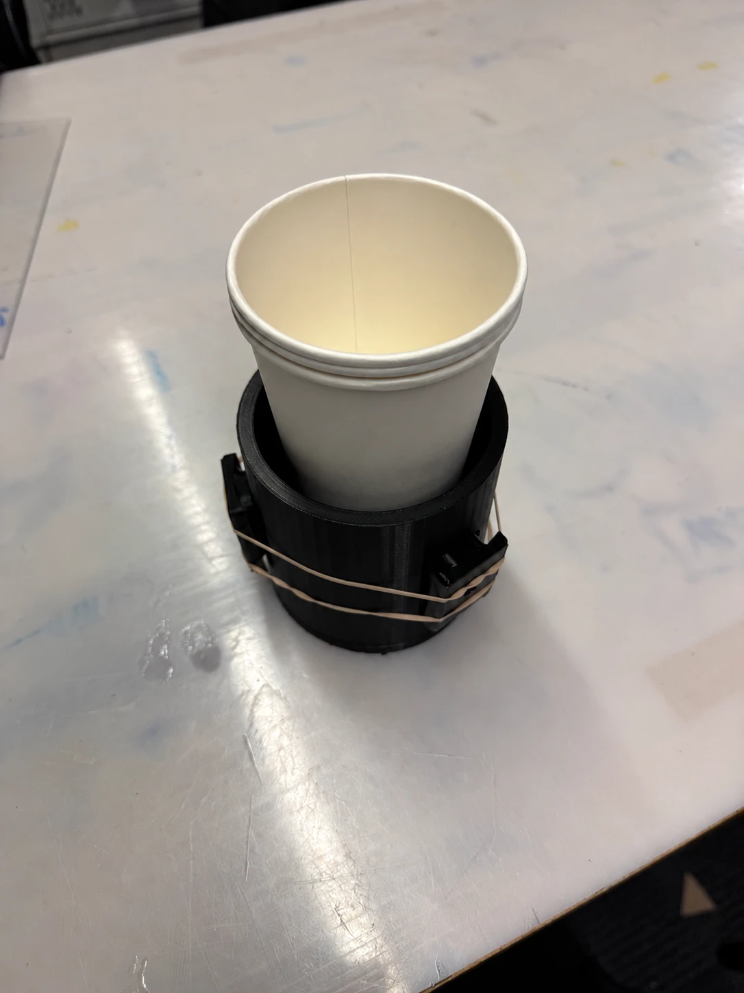 Paper cup inside a black printed cupholder with elastic bands around its outer sections, resting on a worn workshop table.