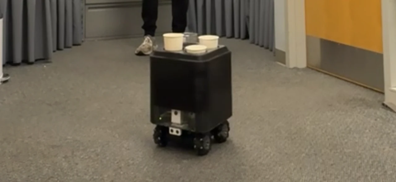 Front view of the robot carrying several cups in a hallway, with its camera and ultrasonic sensor visible below the body.