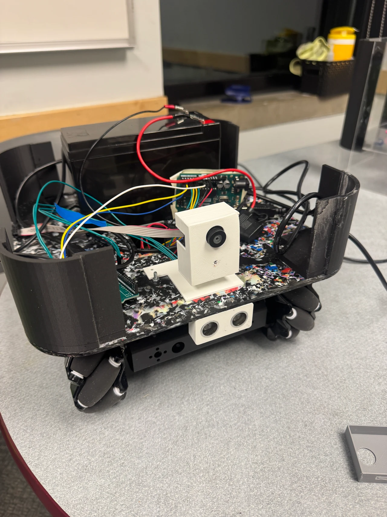 Open robot chassis showing wiring, electronics, a white printed camera enclosure, front ultrasonic sensor, and omnidirectional wheels.